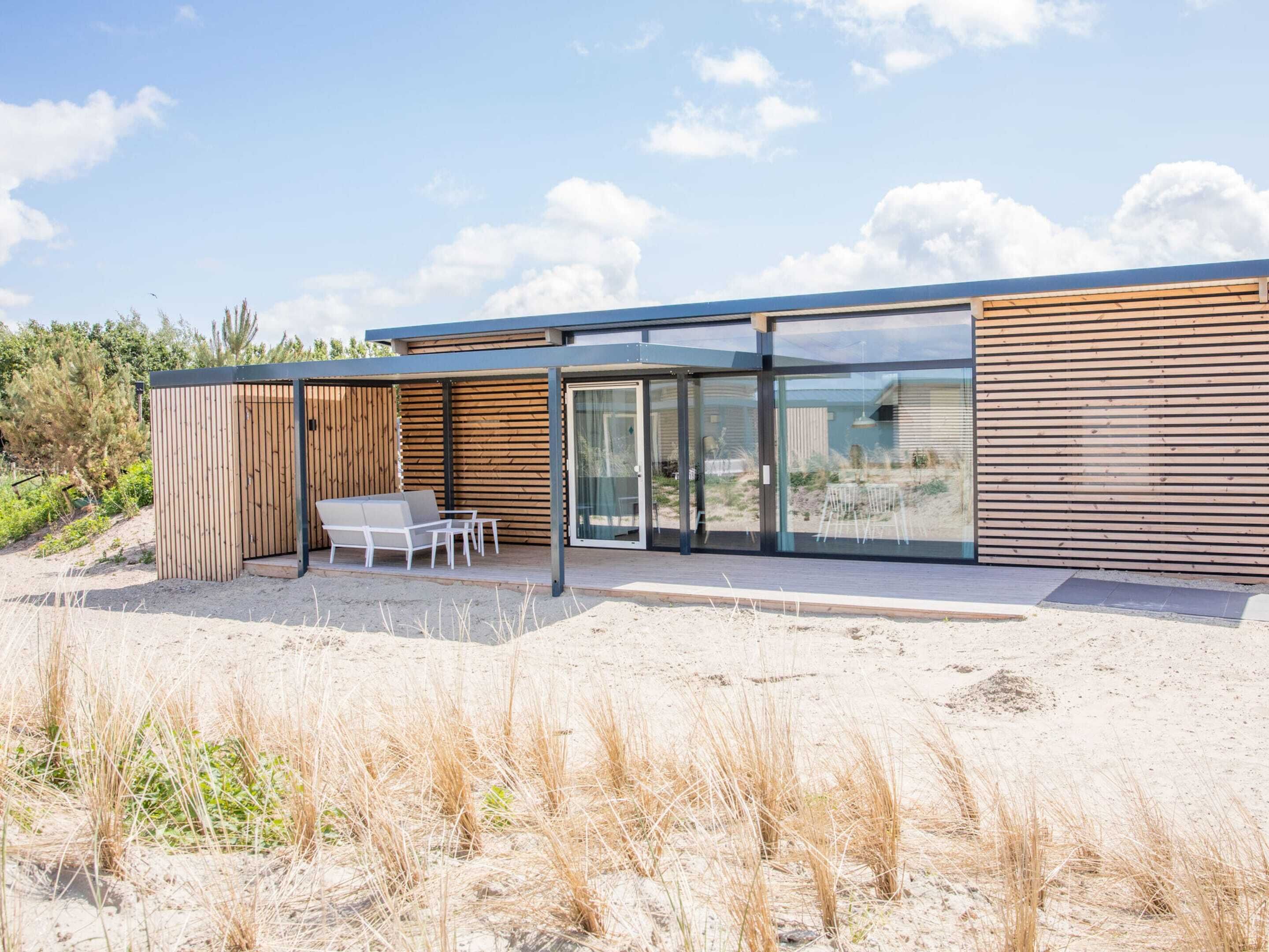 A modern beach house with a glass-enclosed patio, wooden exterior, and a seating area with a table and chairs.