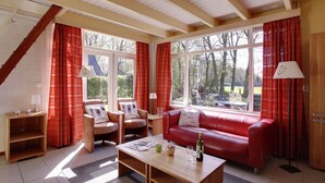 Un salon chaleureux avec un canapé rouge, deux fauteuils, une table basse en bois et un téléviseur. De grandes fenêtres offrent une vue sur les arbres et un jardin.