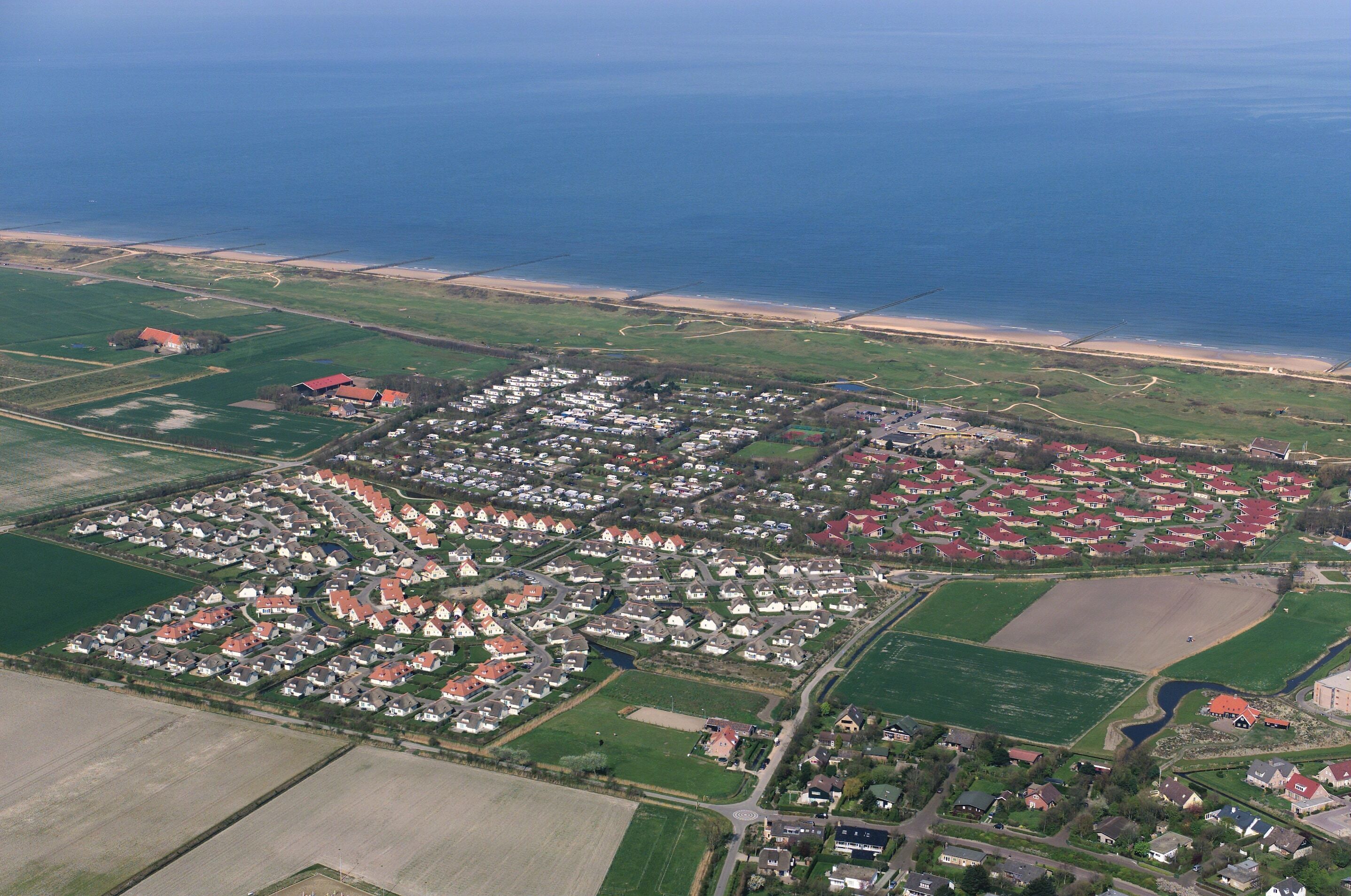Vue aérienne d’une zone résidentielle côtière, avec des maisons, des champs verdoyants et une plage.