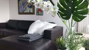 A dark leather sofa with a white pillow and folded grey blanket, a large green monstera plant, and two smaller potted plants on a table. - Unique Holiday Home in Bergen aan Zee With Terrace (Bergen aan Zee)