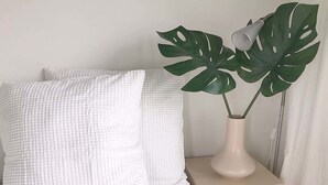 A bed with white textured pillows, a beige bedside table with a white vase containing a plant, and a white curtain in the background. - Unique Holiday Home in Bergen aan Zee With Terrace (Bergen aan Zee)