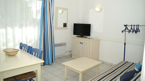 Um quarto com televisão sobre um móvel, uma mesa de jantar com cadeiras, uma mesinha, um sofá com manta listrada e um cabideiro.