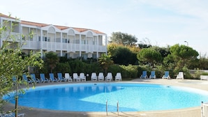 Uma piscina com espreguiçadeiras, um edifício com varandas e vegetação.