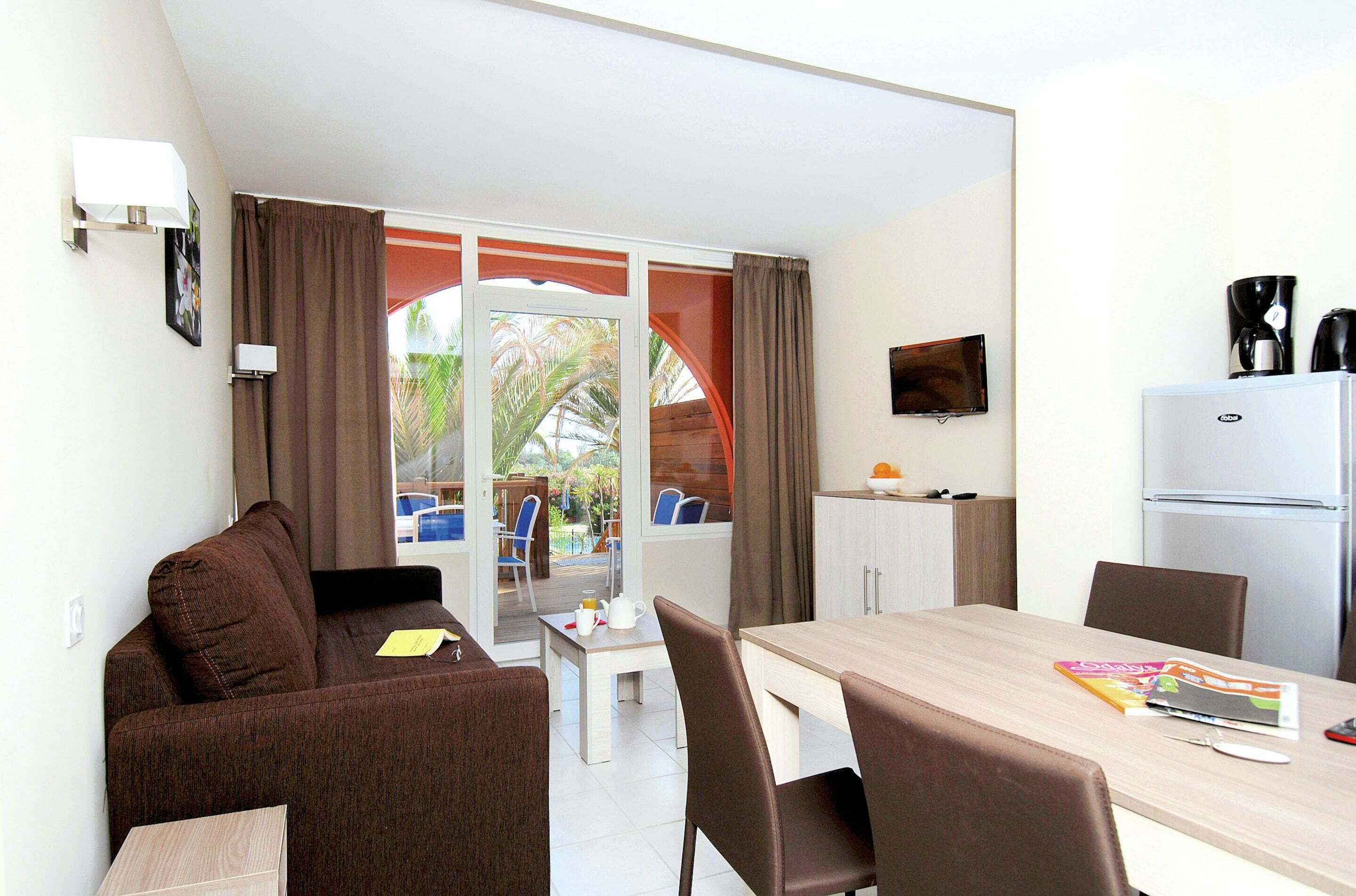 A room with a sofa, a dining table with chairs, a refrigerator, a television, and a window with curtains.