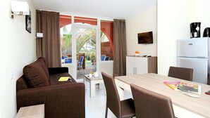 Una habitación con sofá, una mesa de comedor con sillas, refrigerador, televisor y ventana con cortinas.