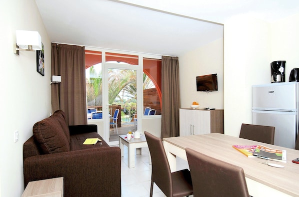 A room with a sofa, a dining table with chairs, a refrigerator, a television, and a window with curtains. - Apartment Near Beach in Lively Resort (Agde)