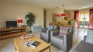 A living room with a TV, wooden entertainment unit, grey sofas, a wooden coffee table, and a dining area with red curtains. - Villa in France With Private Garden (Lanzac)