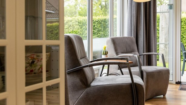 A modern living room with two grey armchairs, a wooden coffee table, and a view of greenery through large windows. - Canal-side Bungalow Stay (Earnewald)
