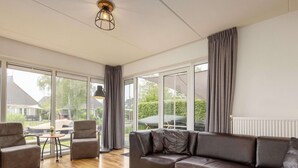 A spacious living room with a large sofa, two round wooden tables, and a view of the outdoors through large windows. - Canal-side Bungalow Stay (Earnewald)
