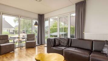 A spacious living room with a large sofa, two round wooden tables, and a view of the outdoors through large windows.