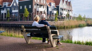 Un couple est assis sur un banc, face à un canal, avec des maisons traditionnelles en arrière-plan.