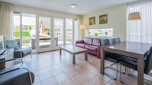 A modern hotel room with a dining area, a red leather sofa, a wooden dining table, and a view of the outside through large sliding glass doors. - Apartment in Waterpark With Private Jetty (Uitgeest)