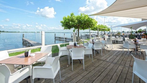 An outdoor dining area with tables and chairs, a wooden deck, and a view of the water. - Apartment in Waterpark With Private Jetty (Uitgeest)
