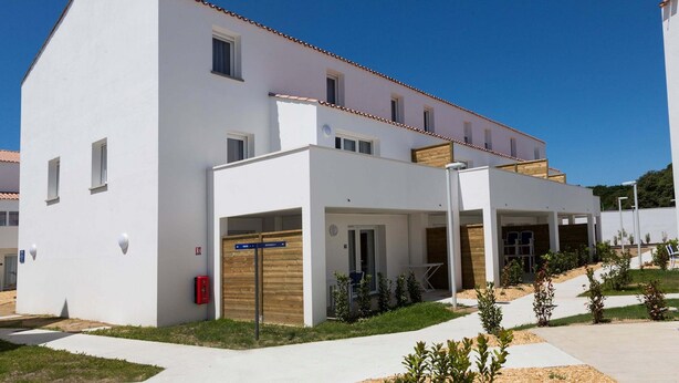 Ein modernes Gebäude mit weißer Fassade, zahlreichen Fenstern und einem überdachten Außenbereich mit Sitzgelegenheiten.