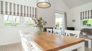 A dining area with a wooden table, white chairs, a hanging light fixture, and a view of greenery outside. - Well-furnished Home With a Porch Near the Sea (Noordwijk)