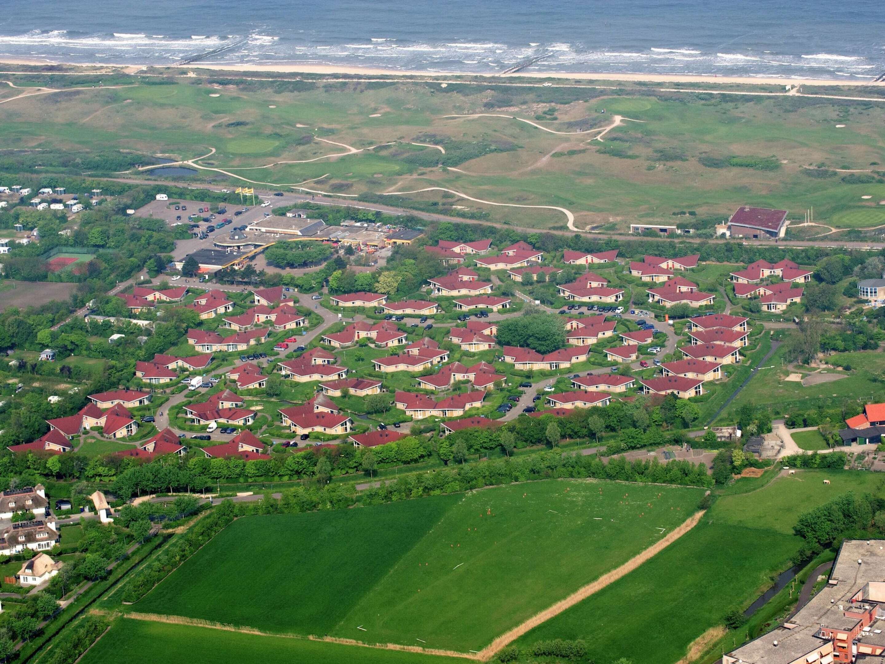 An aerial view of a residential area with houses arranged in a grid pattern, surrounded by green fields and a coastline in the background.