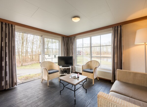 Ein Zimmer mit Fernseher, zwei Korbstühlen, einem Sofa, einem Tisch und großen Fenstern mit Vorhängen.