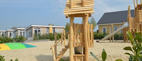 A wooden play structure in a sandy area with a building in the background.