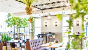 A modern cafe with multiple seating areas, including tables and a sofa, surrounded by potted plants and hanging greenery.