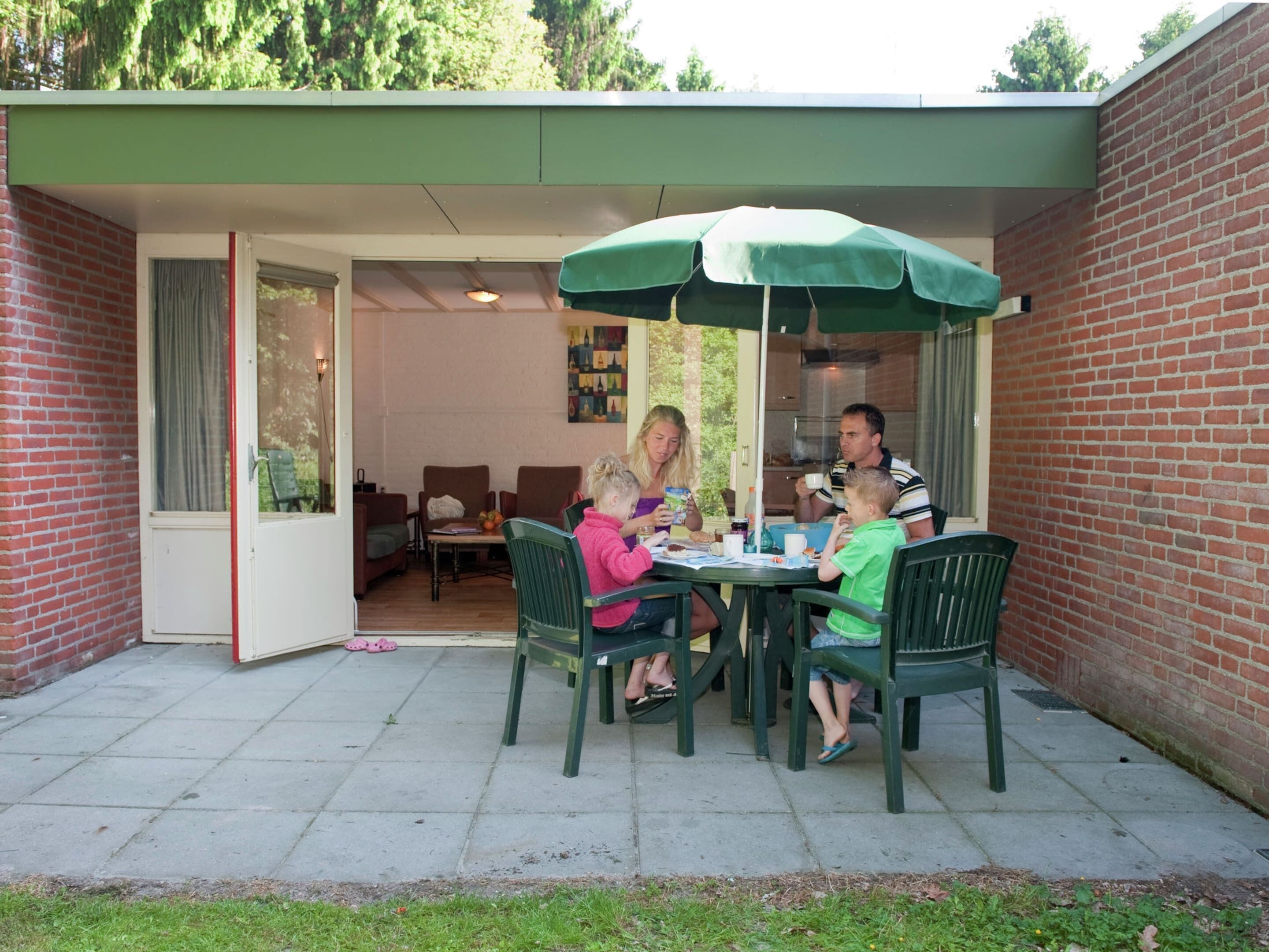 A family dining at an outdoor patio with a green umbrella, brick walls, and a view of the interior dining area.