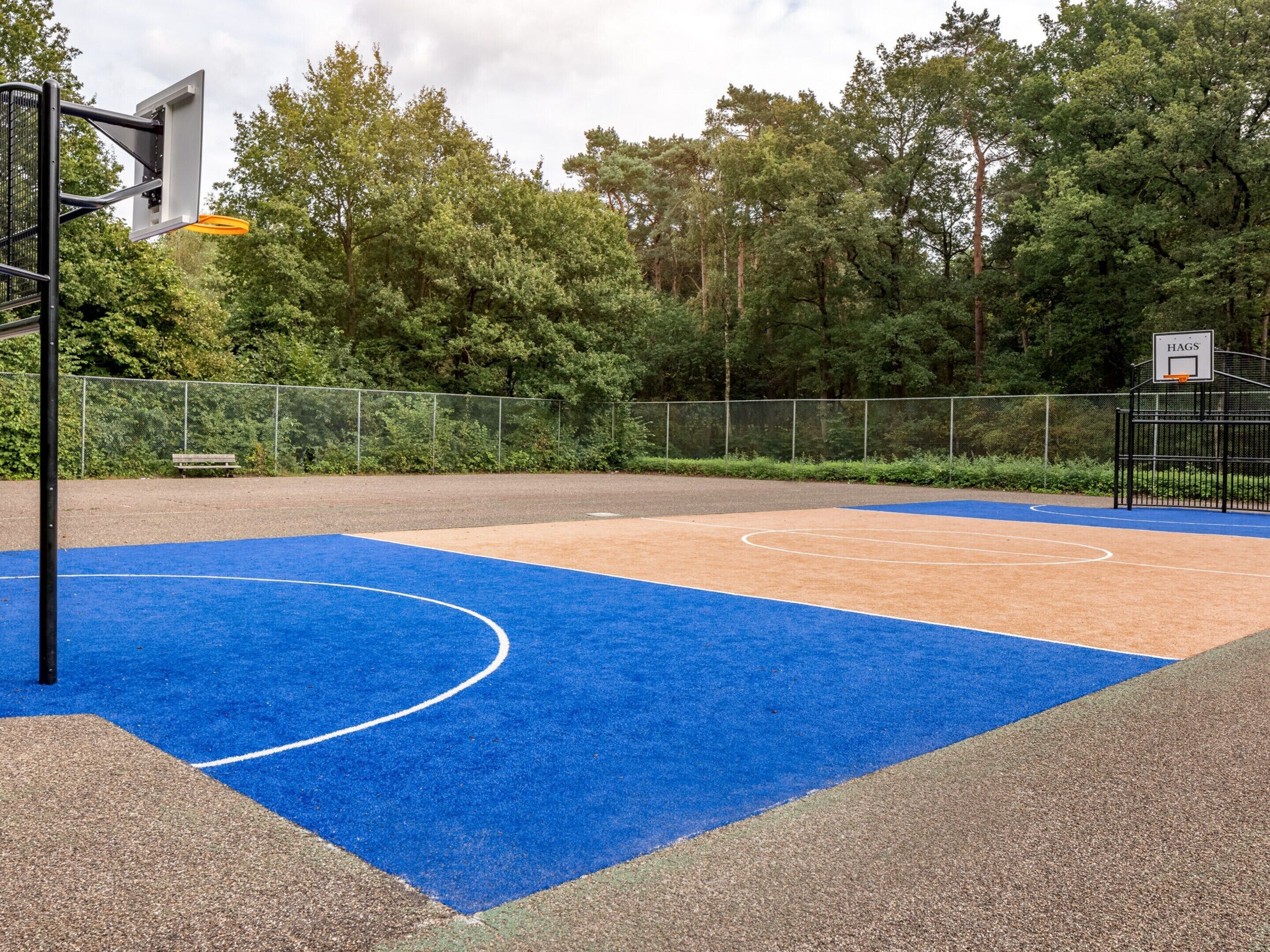 Un terrain de basket en plein air, doté d’un revêtement bleu et beige, entouré d’une clôture en métal et d’arbres.