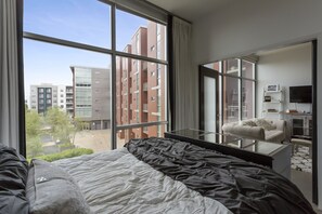 Una camera d'hotel moderna con una grande finestra, un letto con copripiumino bianco e nero, un tavolino da caffè in vetro, un divano e una televisione a schermo piatto.