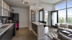 A modern kitchen with stainless steel appliances, a dining area with a white island, and a large window offering a city view. - East Nashville Condo | Rooftop + Parking | 30+ Days (Nashville)