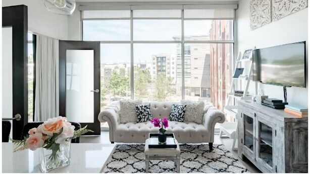 A modern living room with a large window, a white sofa with patterned cushions, a coffee table, and a television on a wooden cabinet. - East Nashville Condo | Rooftop + Parking | 30+ Days (Nashville)