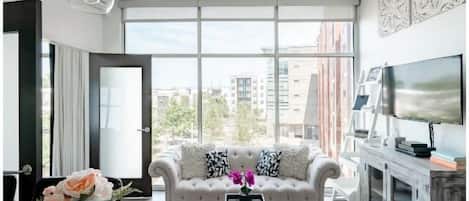 A modern living room with a large window, a white sofa with patterned cushions, a coffee table, and a television on a wooden cabinet.