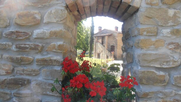 A stone archway with red flowers in the foreground and a building in the background. - Belvilla by OYO Il Gelso (Modigliana)