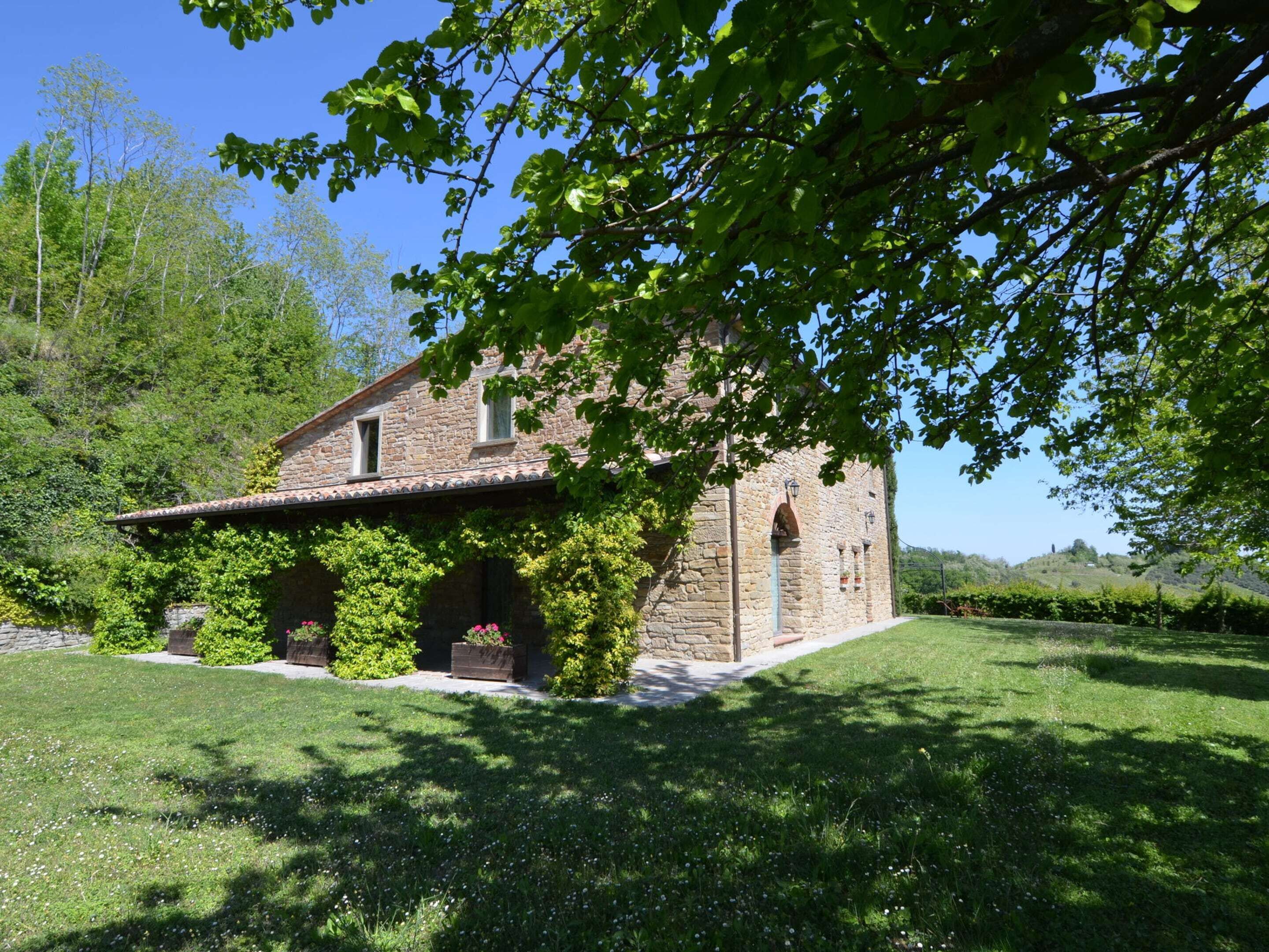 A stone house with a tiled roof, surrounded by a well-maintained lawn and trees. - Belvilla by OYO Il Gelso (Modigliana)