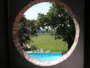 Une vue à travers une fenêtre circulaire donnant sur une piscine entourée d’une pelouse verdoyante et d’arbres.