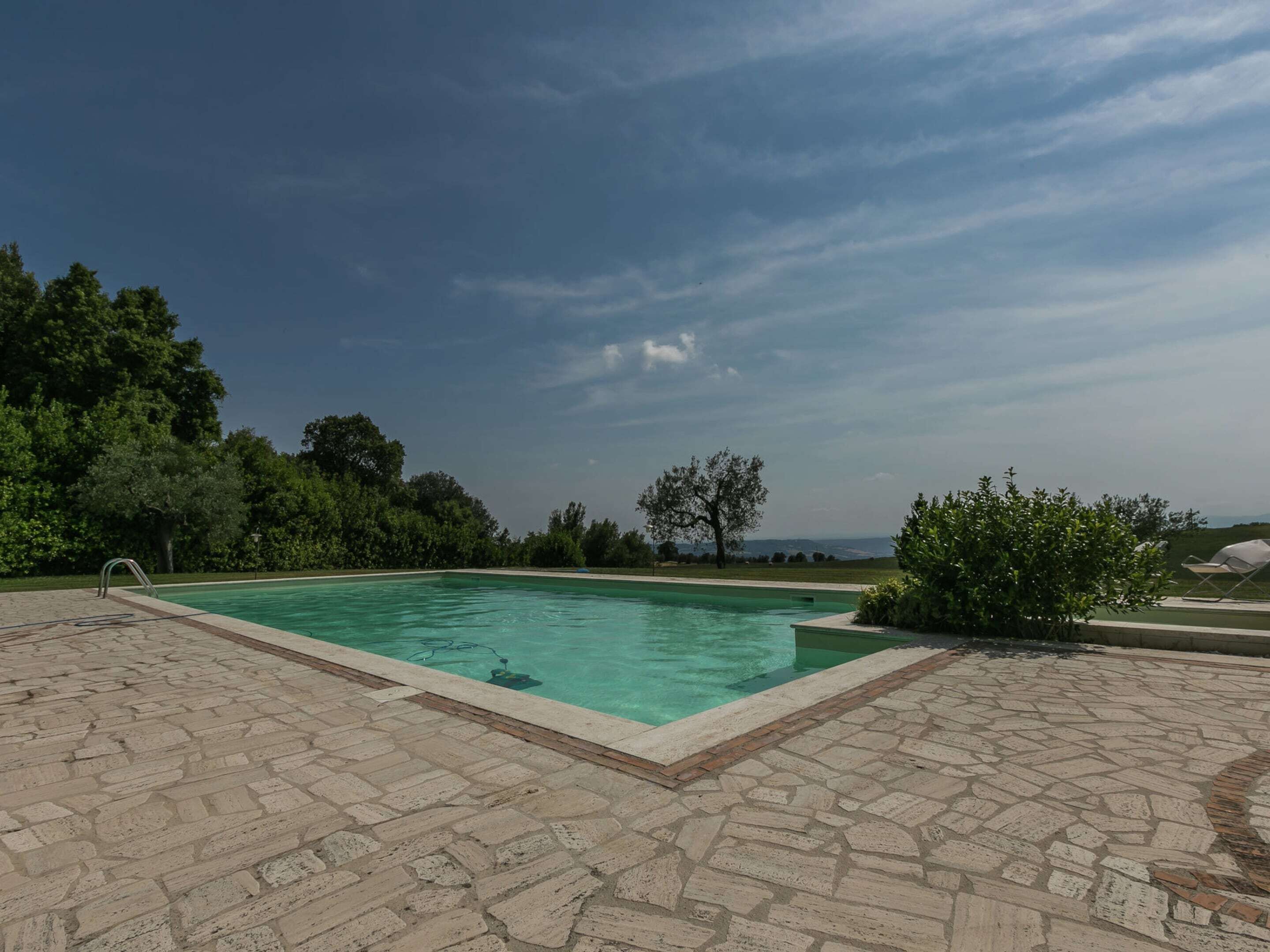 Une piscine entourée d’arbres, avec une terrasse dallée de pierres et un ciel dégagé.
