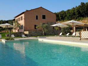 A pool area with lounge chairs and umbrellas, a stone wall, and a building with a flat roof. - Luxury, Modern Apartment With Pool and Stunning Views (Narni)