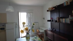 Una cocina con refrigerador, una mesa de comedor con sillas, un florero con flores y una estantería con libros.