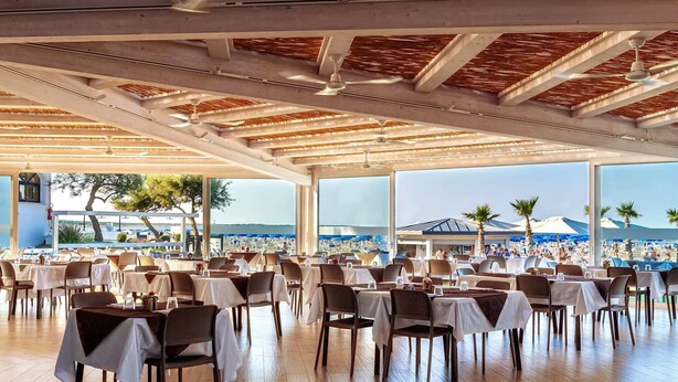 An outdoor dining area with tables and chairs, large windows, and a view of a beach and mountains.
