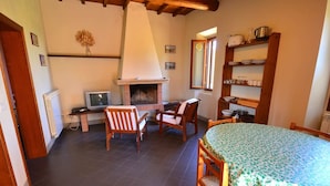 A cozy kitchen with a fireplace, wooden chairs, a dining table with a floral tablecloth, and a television on a side table. - Scenic Apartment With Pool (Figline e Incisa Valdarno)