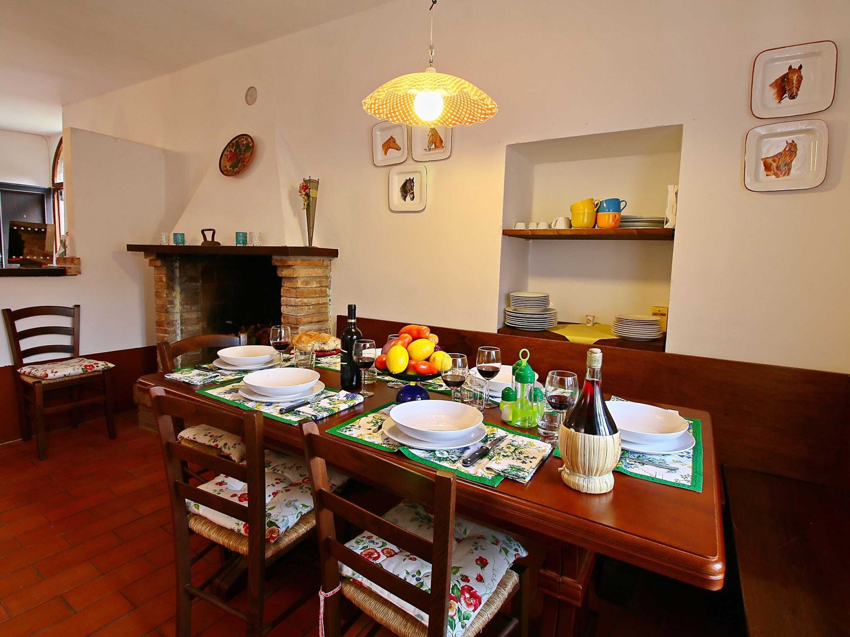 A dining room with a wooden table set for four, surrounded by chairs with floral cushions. A fireplace is visible in the background, and the room has a cozy atmosphere with a hanging lamp above the table.