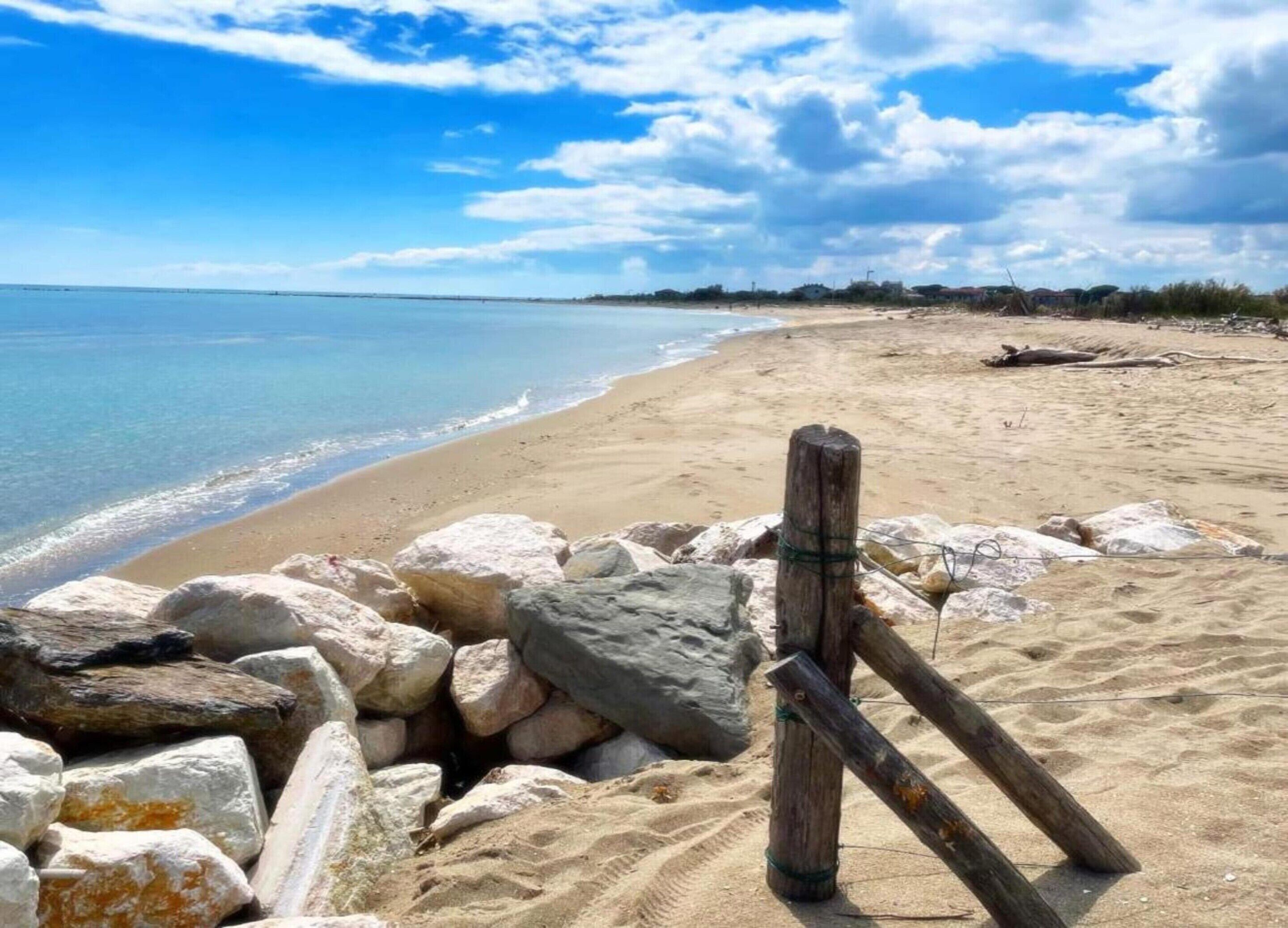A beach with a wooden fence and large rocks in the foreground, a clear sky, and calm sea.