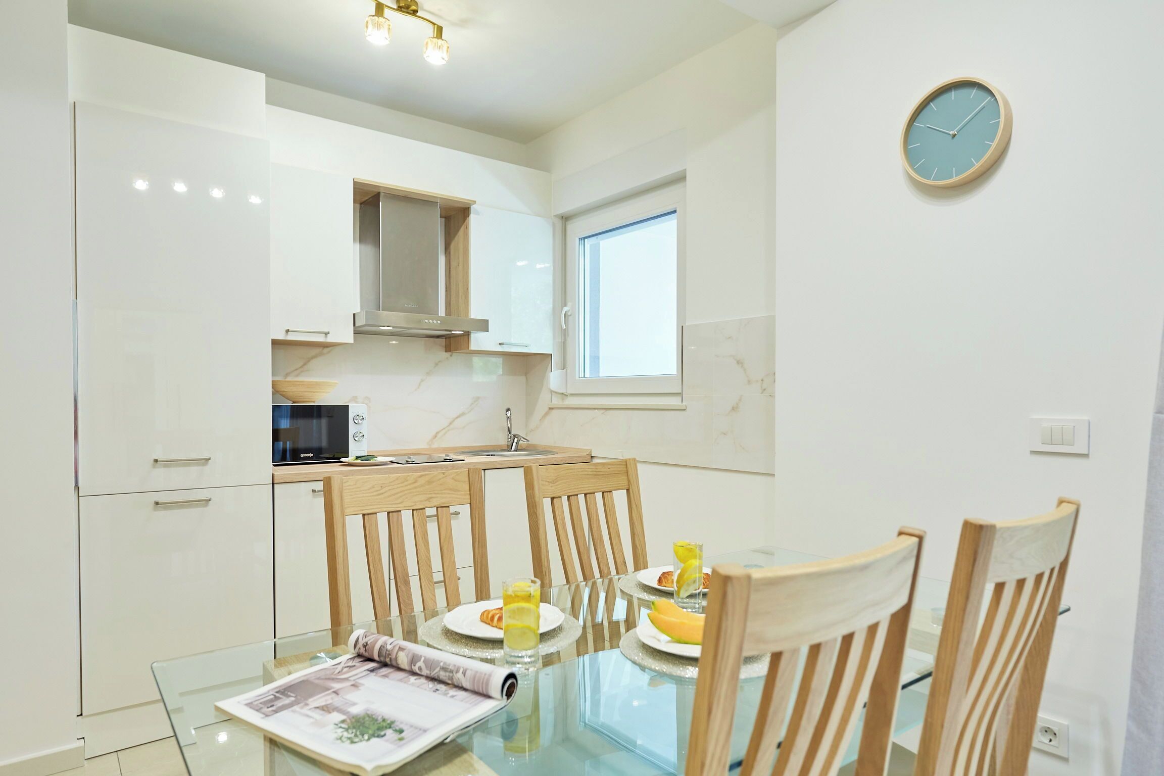Een moderne keuken met witte kasten, een magnetron en een raam. Er staat een eettafel met stoelen en een klok hangt aan de muur.