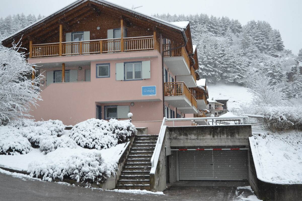Un edificio de varios pisos con un balcón de madera, un letrero que dice 'Le P'tit Bateau', y un paisaje cubierto de nieve.