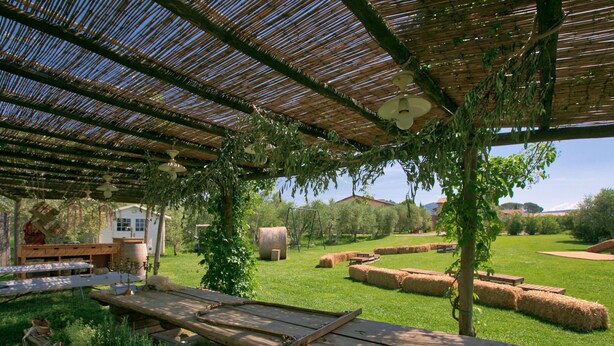 An outdoor patio area with a wooden table, a pergola with a ceiling fan, hay bales, and a grassy field with trees in the background. - Tuscan Stay w/ Jacuzzi (Grosseto)