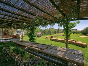 An outdoor patio area with a wooden table, a pergola with a ceiling fan, hay bales, and a grassy field with trees in the background.
