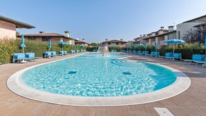 Une piscine dotée d’un élément décoratif central, entourée de chaises longues et de parasols, avec des bâtiments en arrière-plan.