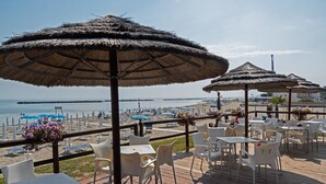 Un restaurant situé en bord de mer, avec une terrasse, des parasols en chaume et une vue sur l’océan.