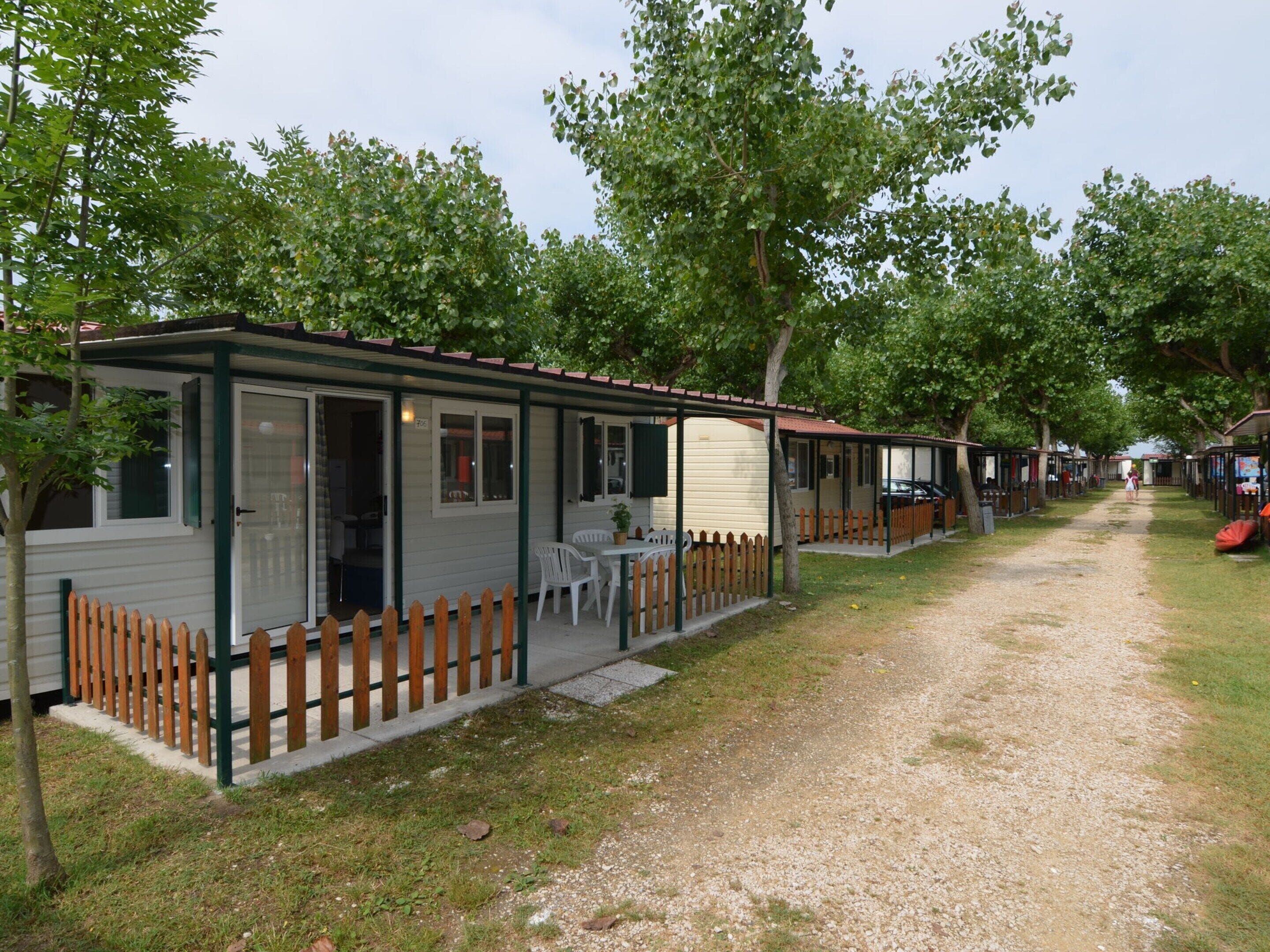 A row of mobile homes with attached porches, each with a small table and chairs, set in a grassy area with trees.