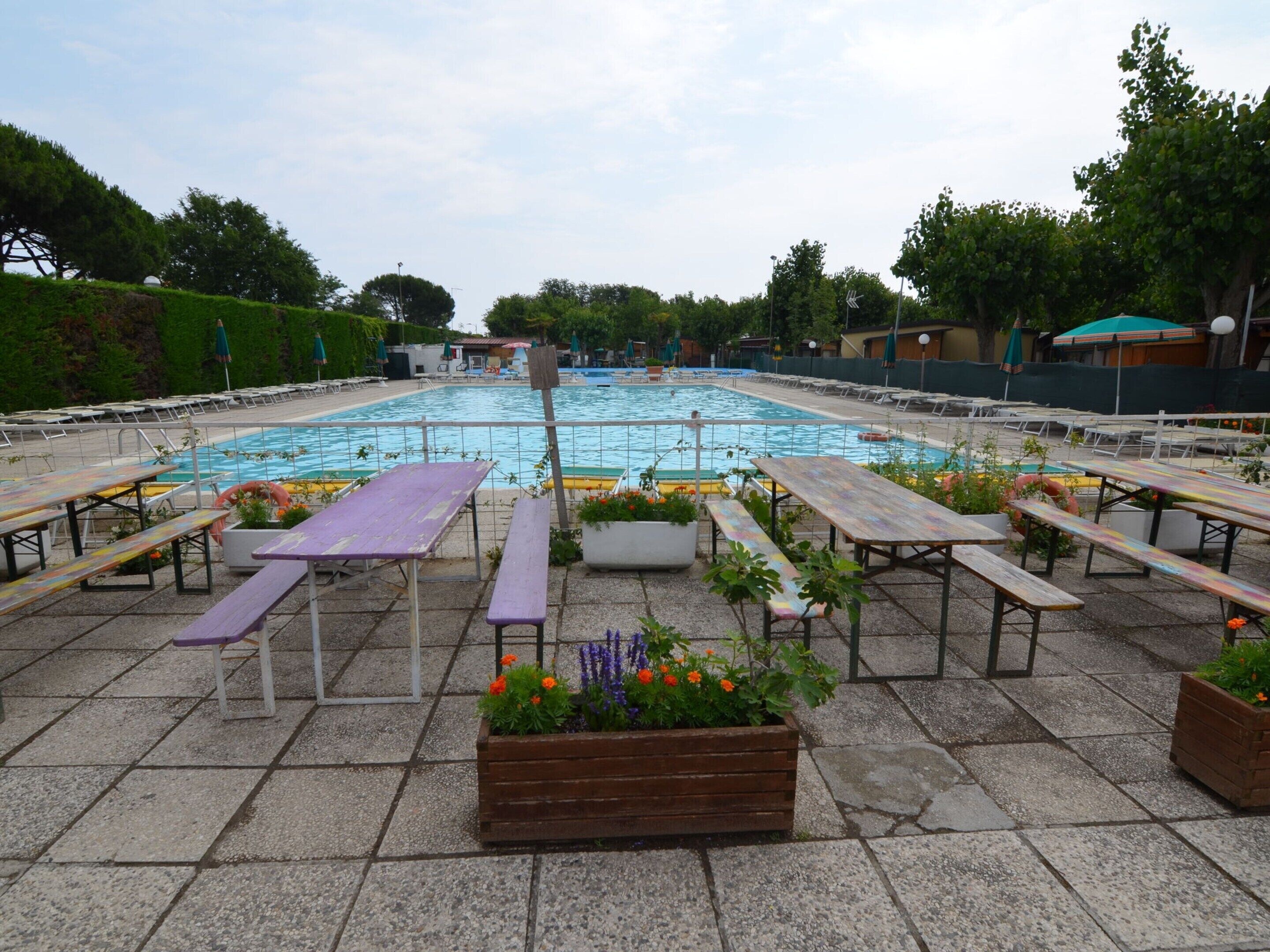 A swimming pool with surrounding picnic tables and benches, surrounded by greenery and trees.