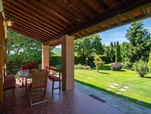 An outdoor patio with a brick floor, wooden furniture, and a view of a well-maintained garden.