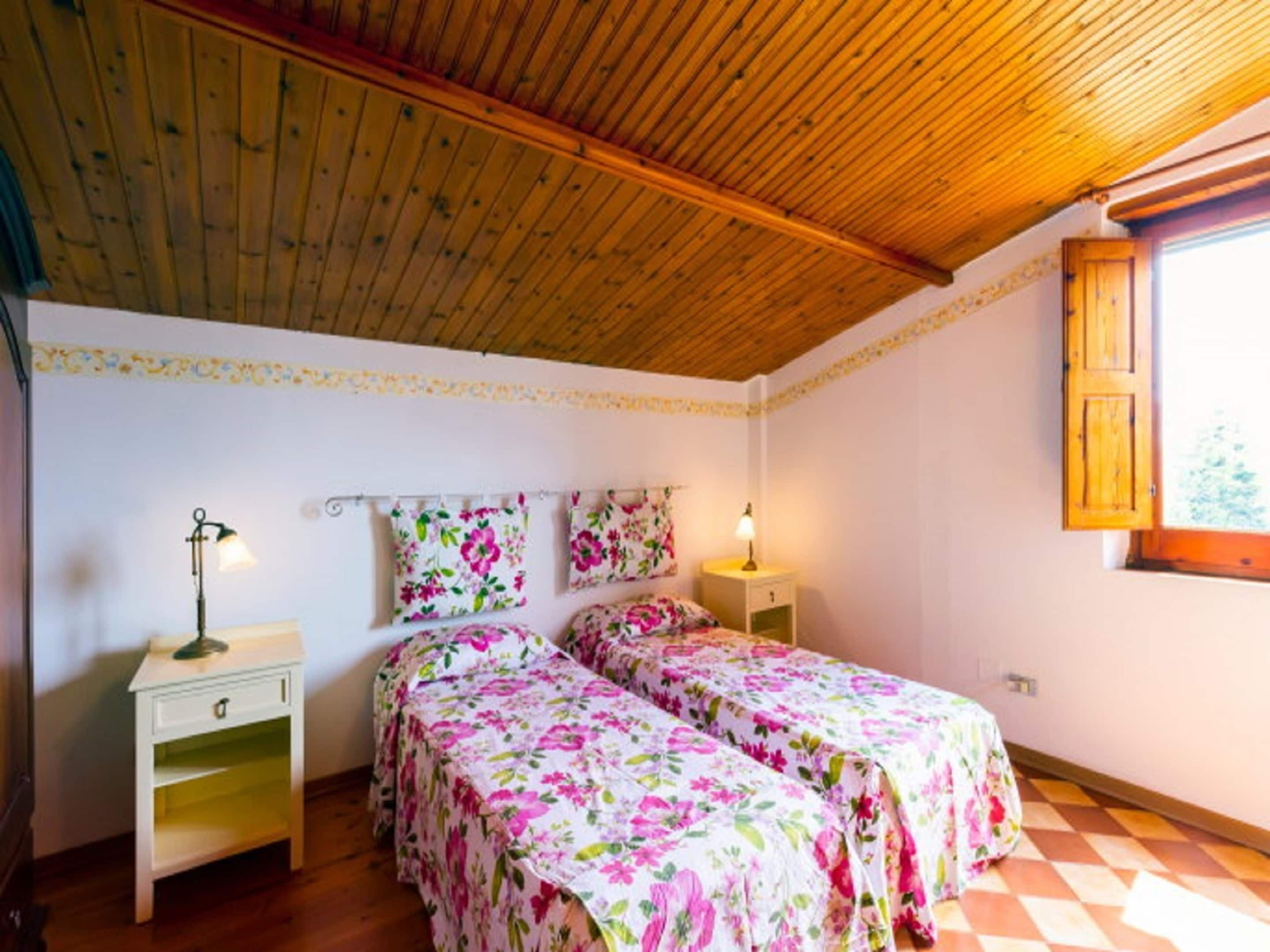 A bedroom with two beds, wooden ceiling, floral bedspreads, bedside tables, and a window with wooden shutters.