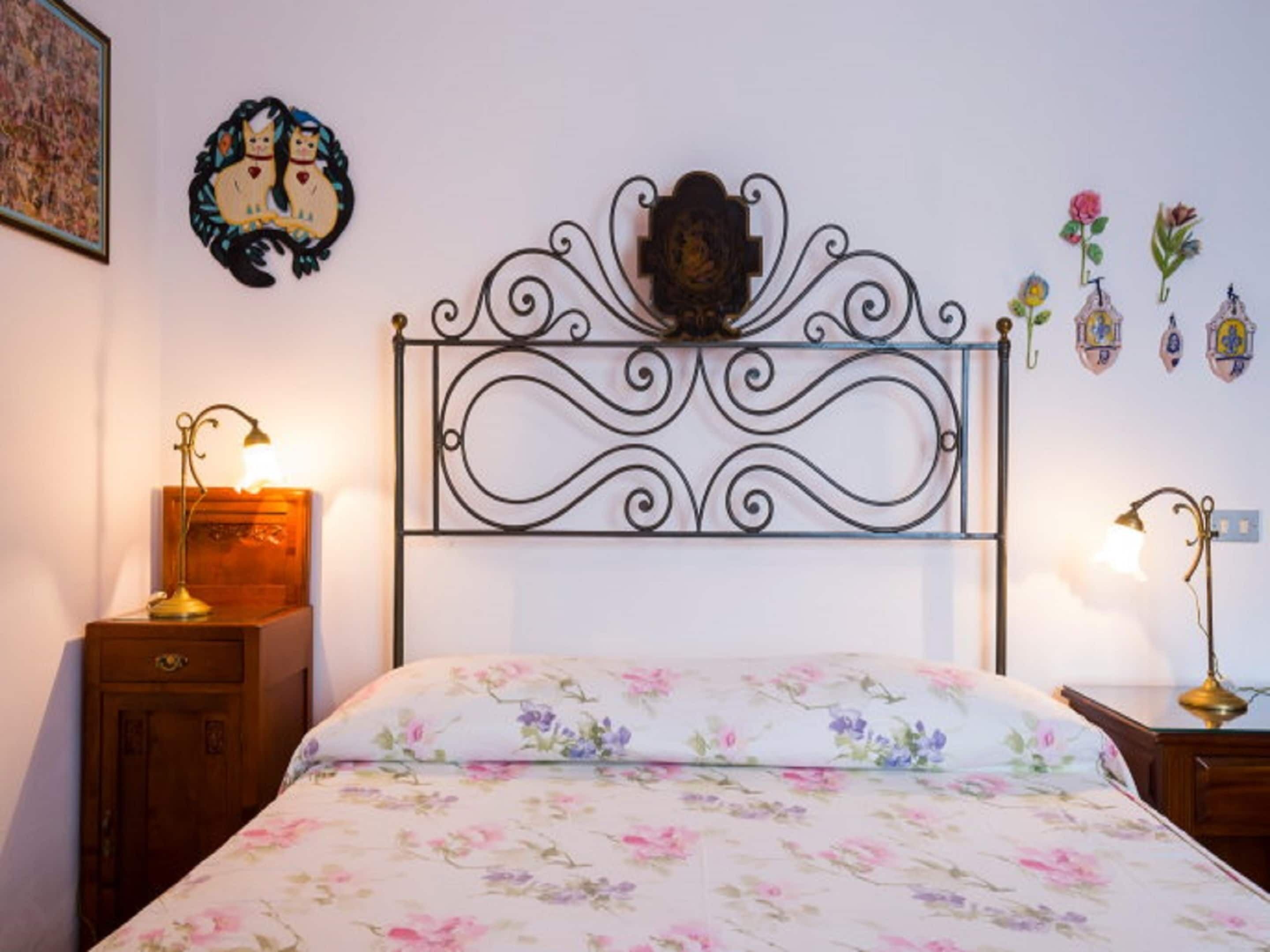A bedroom with a floral bedspread, a decorative headboard, two bedside tables with lamps, and wall decorations including a cat illustration and flower patterns.